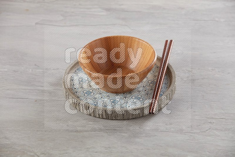 brown wood round bowl on top of multi color round ceramic plate and chopsticks, on grey textured countertop