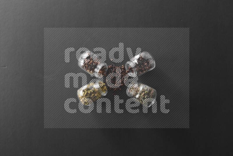 4 glass spice jars full of cloves, cardamom, basil and star anise on black flooring