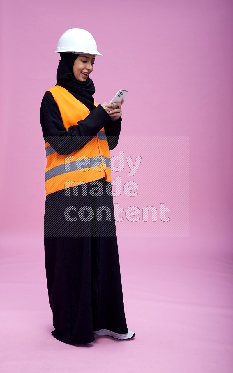 Saudi woman wearing Abaya with engineer vest and helmet standing texting on phone on pink background