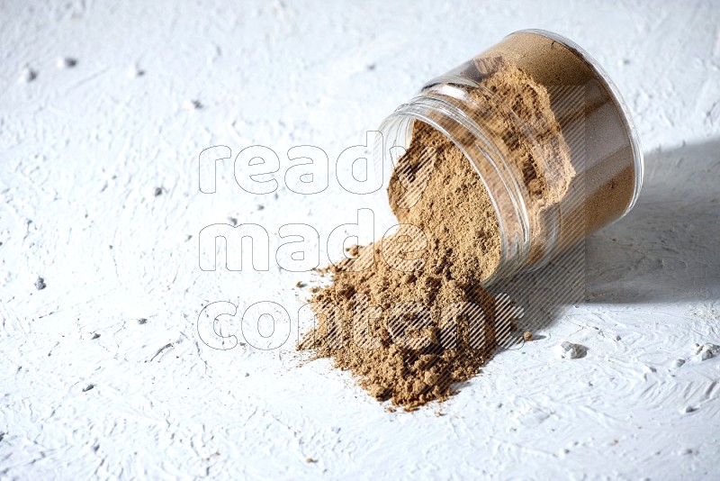 A flipped glass jar full of allspice powder and powder spilled out of it on a textured white flooring