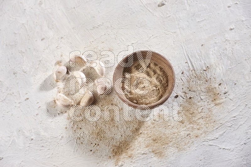 A wooden bowl full of garlic powder and beside it garlic cloves on a textured white flooring in different angles