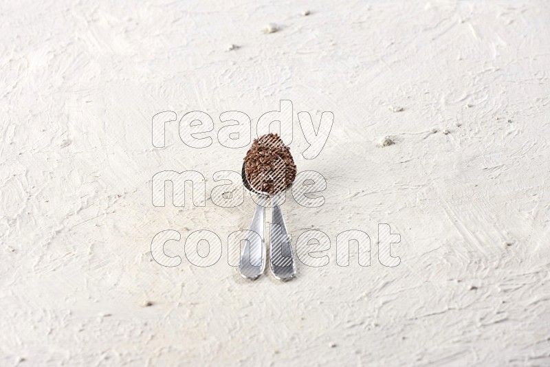 2 metal spoons full of flax seeds on a textured white flooring