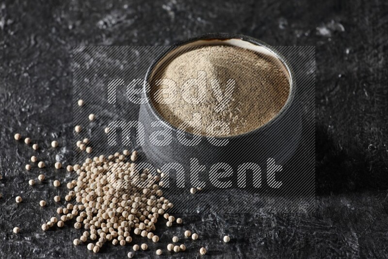 Black pottery bowl full of white pepper powder with white pepper beads on textured black flooring