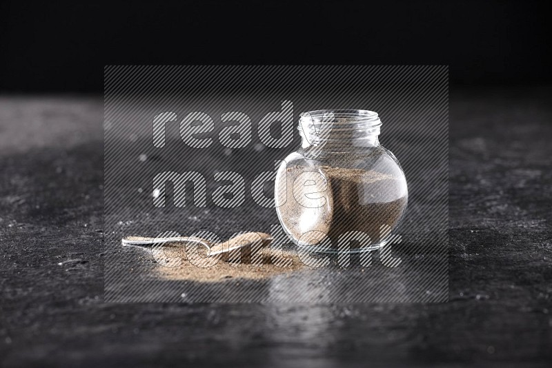 A glass spice jar full of black pepper powder and a metal spoon full of powder on textured black flooring