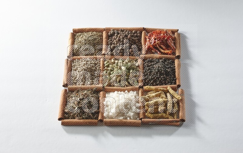 9 مربعات من أعواد القرفة ممتلئة بالهيل في المنتصف محاطة بأصابع الكركم والملح والريحان المجفف والفلفل الأبيض والكمون وبهار الفلفل الحلو والفلفل الحار المجفف والفلفل الأسود على أرضية بيضاء