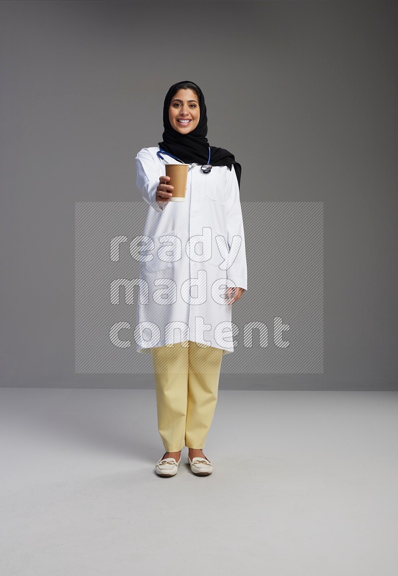 Saudi woman wearing lab coat with stethoscope standing holding paper cup on Gray background