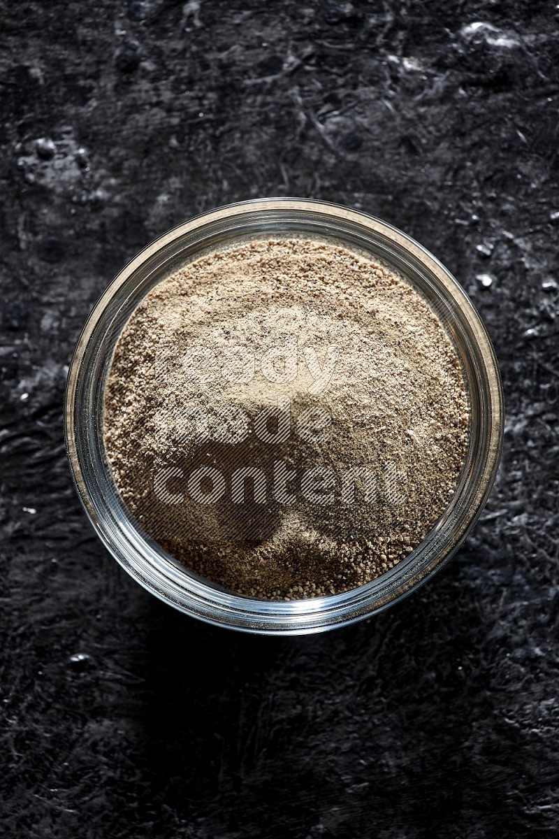 A glass bowl full of white pepper powder on textured black flooring