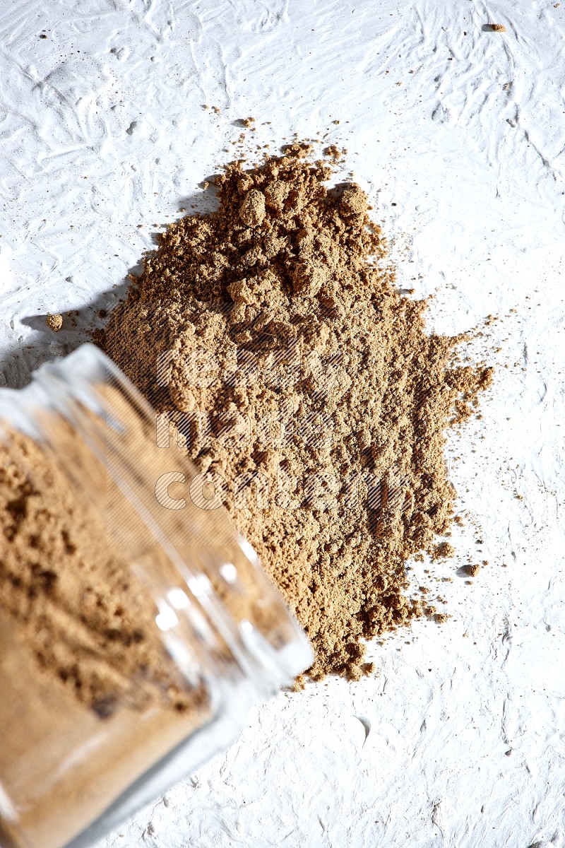 A flipped glass jar full of allspice powder and powder spilled out of it on a textured white flooring