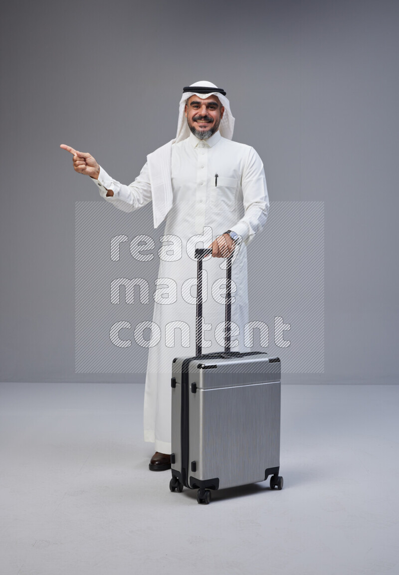 Saudi man wearing Thob and white Shomag standing holding Travel bag on Gray background