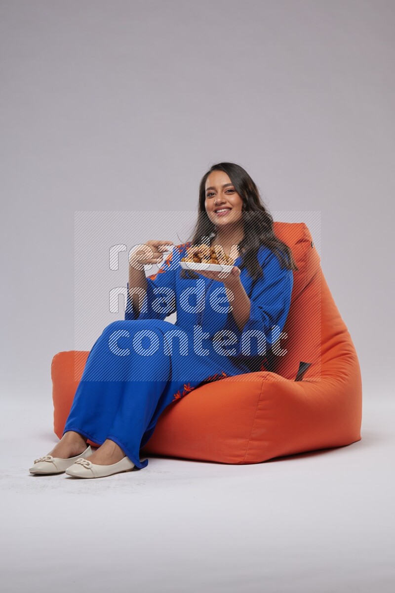 A Woman sitting on an orange beanbag wearing Jalabeya holding a plate of dates