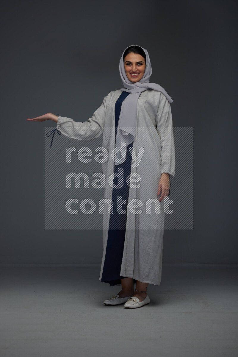 A Saudi woman wearing a light gray Abaya and head scarf standing and pointing in different directions eye level on a grey background