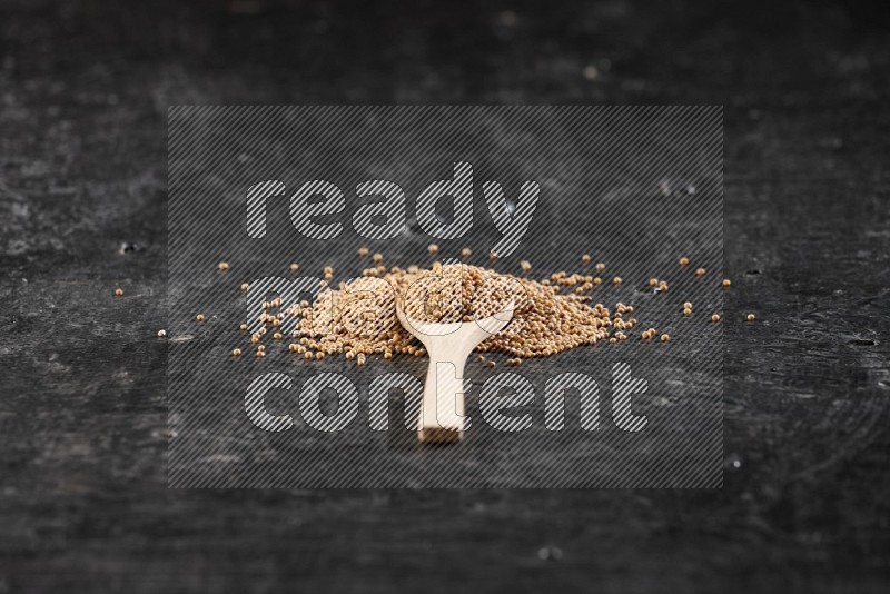 A wooden spoon full of mustard seeds on a textured black flooring