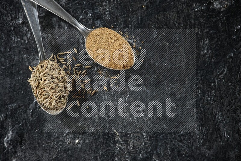 ملعقتان معدنيتان مملوءتان بحبوب الكمون وبودرة الكمون على أرضية سوداء