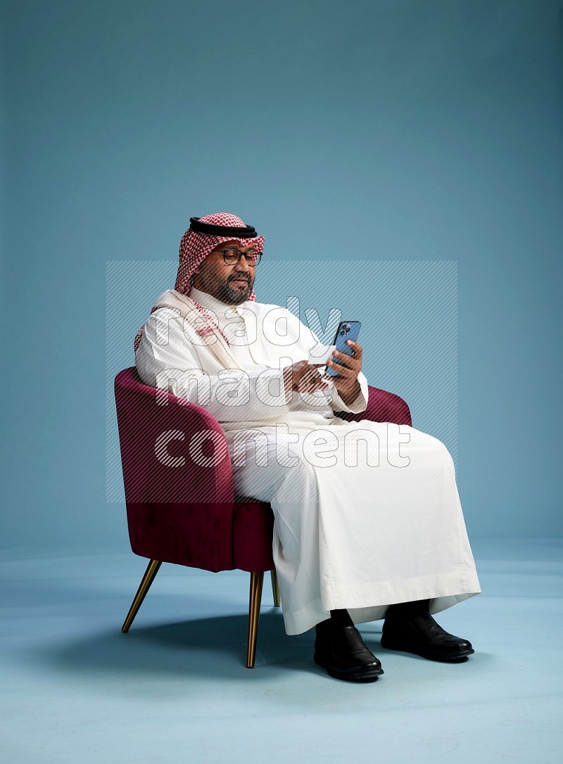 Saudi Man with shimag sitting on chair holding ATM card while talking on phone on blue background