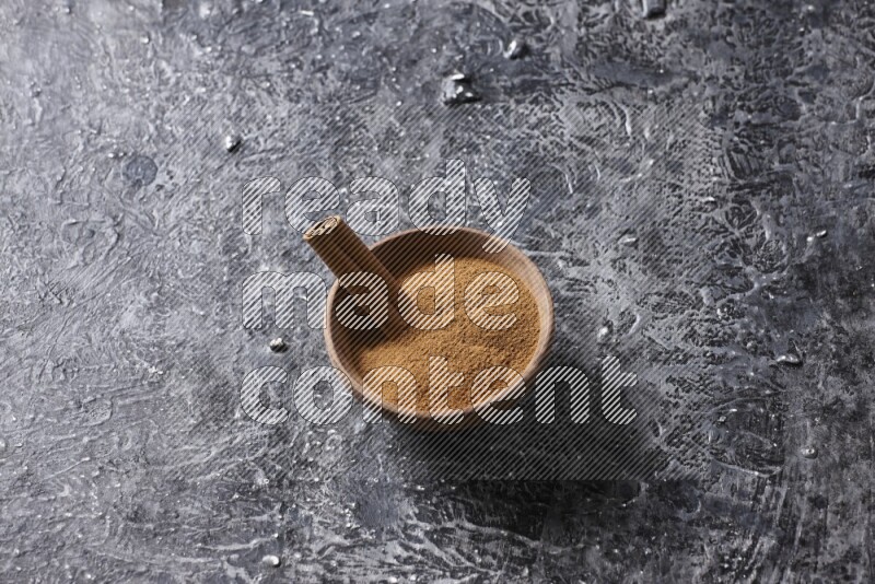 Wooden bowl full of cinnamon powder and a cinnamon stick on a textured black background