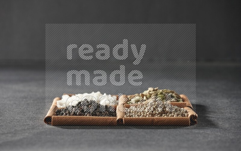 4 مربعات من عيدان القرفة ممتلئة بحبوب الفلفل الأسود والفلفل الأبيض وحب الهيل والملح الأبيض على أرضية سوداء