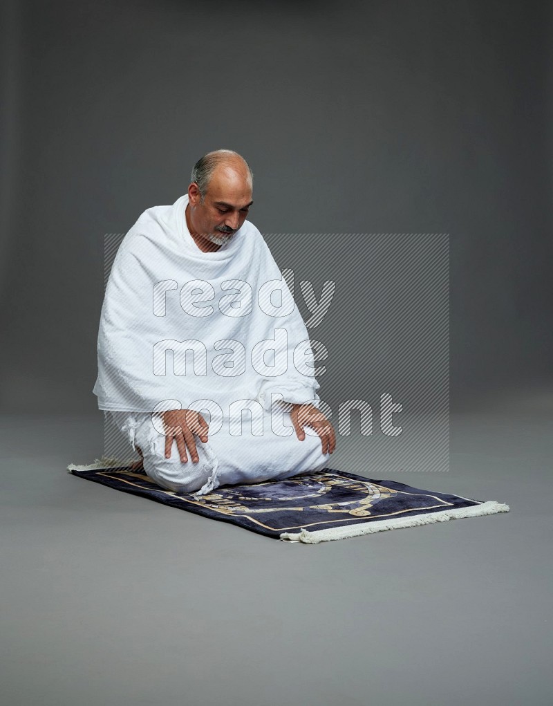 A man wearing Ehram sitting on prayer mat on gray background