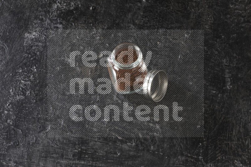 A glass spice jar full of cloves powder on textured black flooring