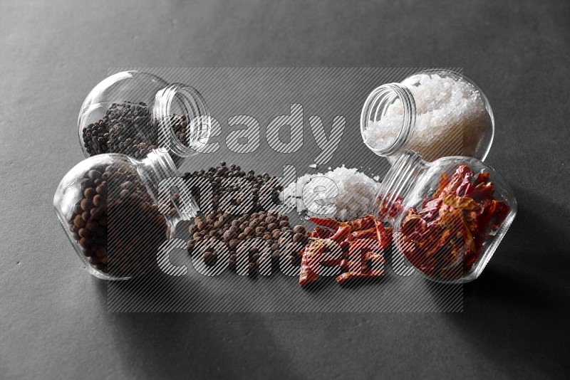 4 glass spice jars full of salt, chili peppers, black peppers and allspice on black flooring