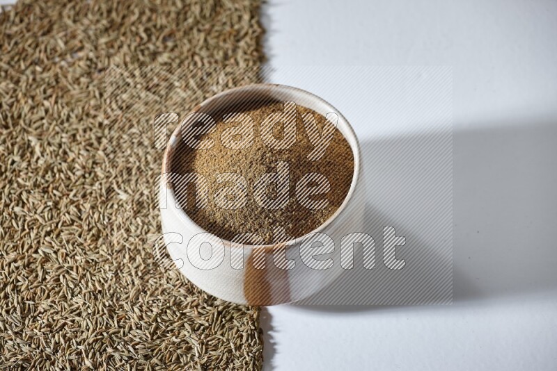 A beige bowl full of cumin powder with cumin seeds beneath it the bowl on a white flooring