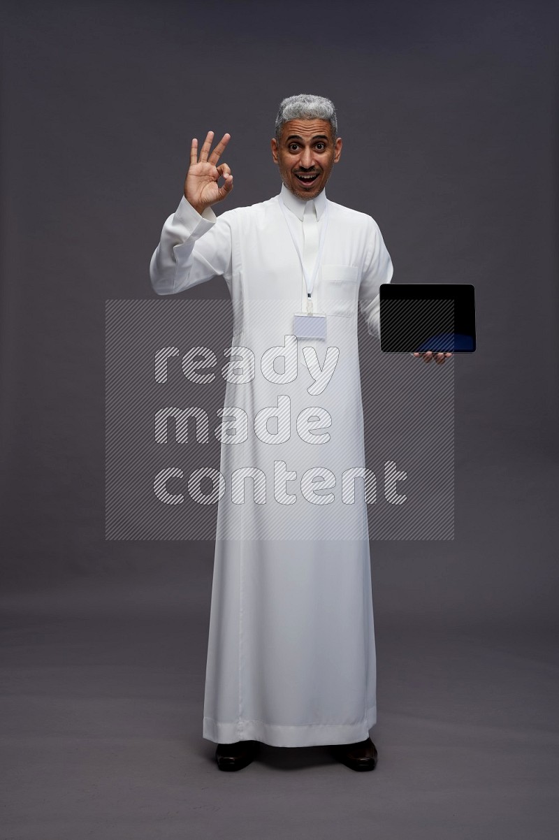 Saudi man wearing thob with neck strap employee badge standing showing tablet to camera on gray background