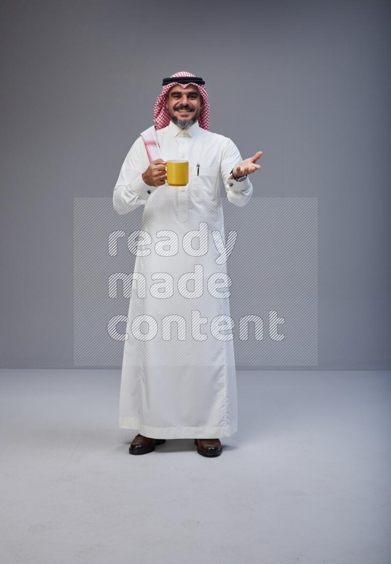 Saudi man Wearing Thob and red Shomag standing holding mug on Gray background
