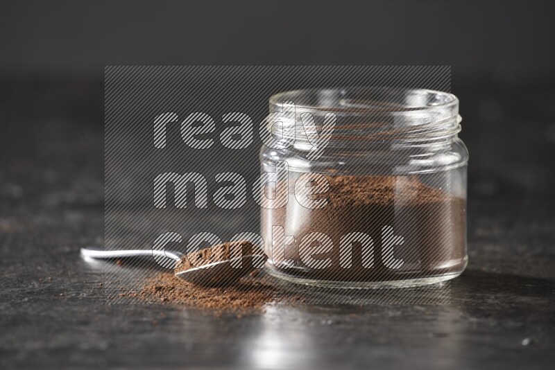 A glass jar full of cloves powder with a metal spoon on a textured black flooring
