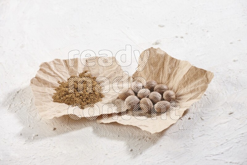 2 crumpled piece of paper full of nutmeg seeds and powder on a textured white flooring