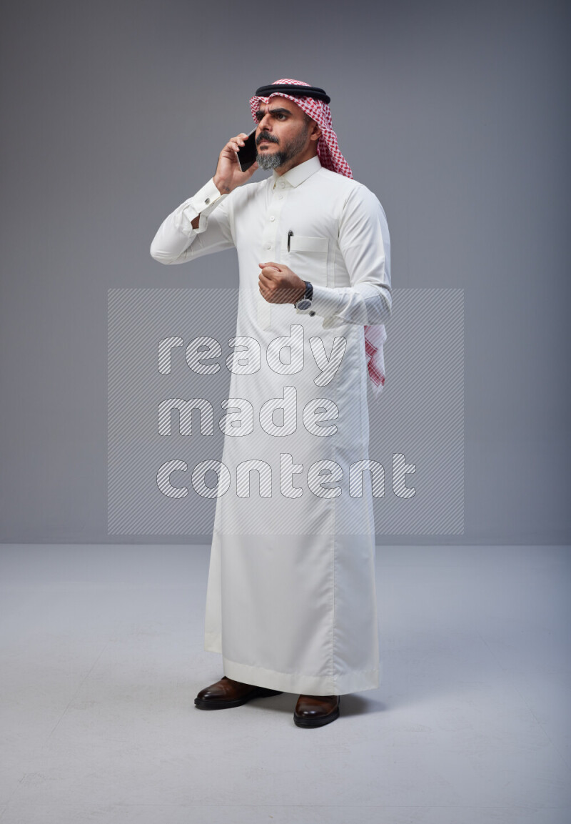 Saudi man Wearing Thob and red Shomag standing talking on phone on Gray background