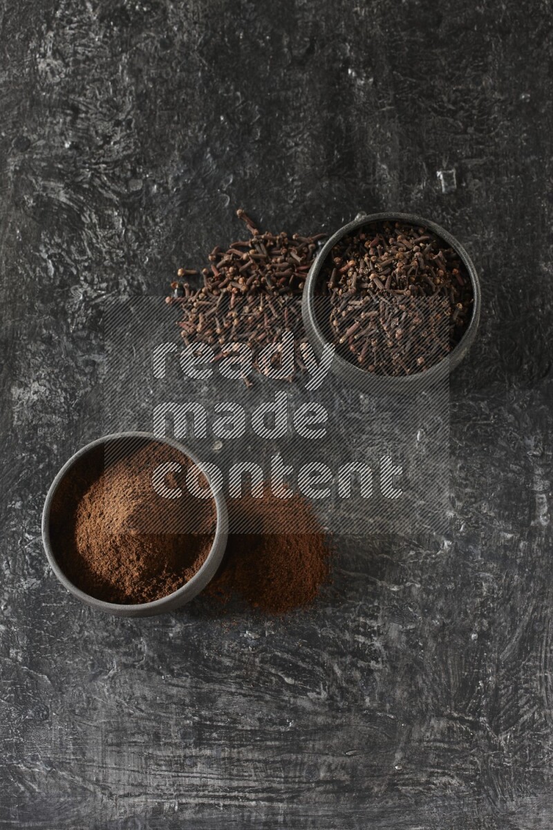 2 Black pottery bowls full of cloves and the other full of cloves powder on textured black flooring