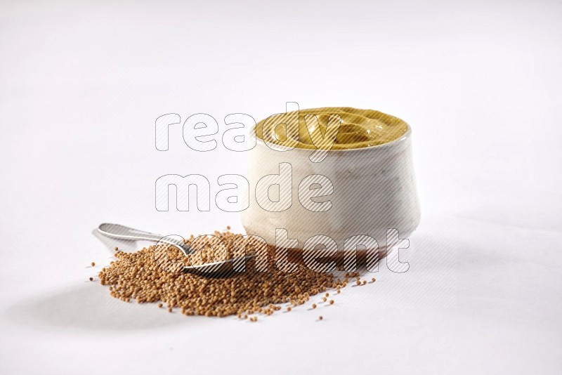 A beige pottery bowl full of mustard paste with mustard seeds underneath with a metal spoon on white flooring in different angles