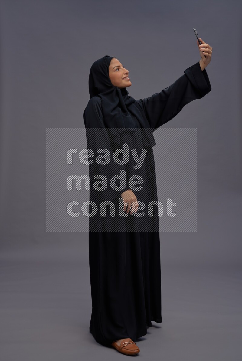 Saudi woman wearing Abaya standing taking selfie on gray background