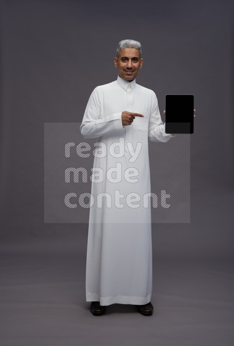 Saudi man wearing thob standing showing tablet to camera on gray background