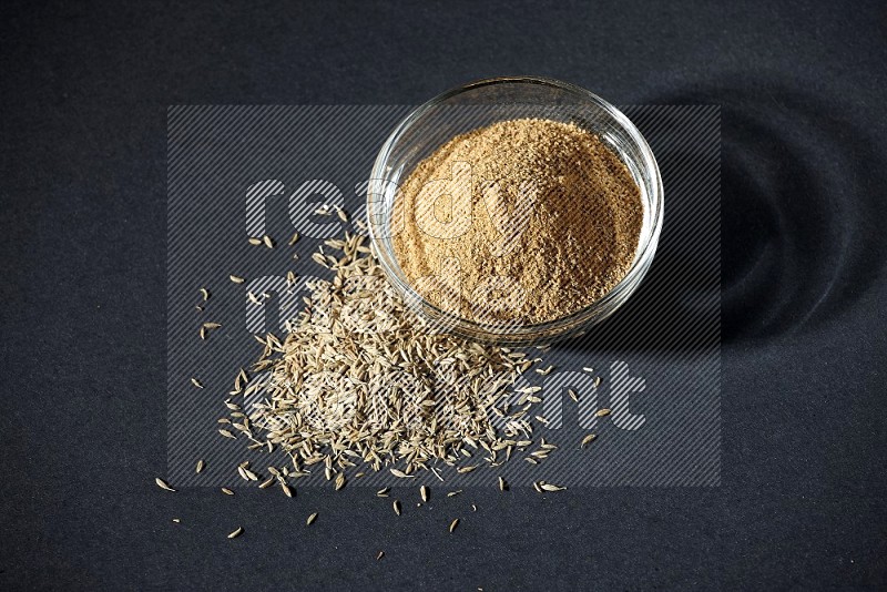 A glass bowl full of cumin powder with cumin seeds beside it on black flooring