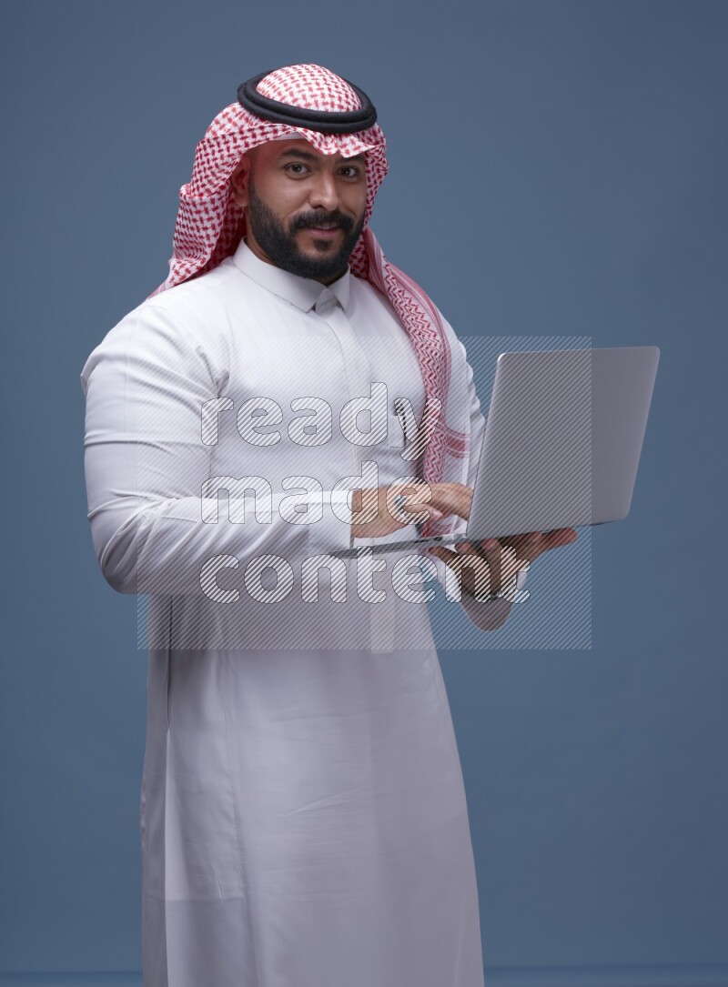 A man standing  with a laptop on Blue Background wearing Saudi Thob and Shomag