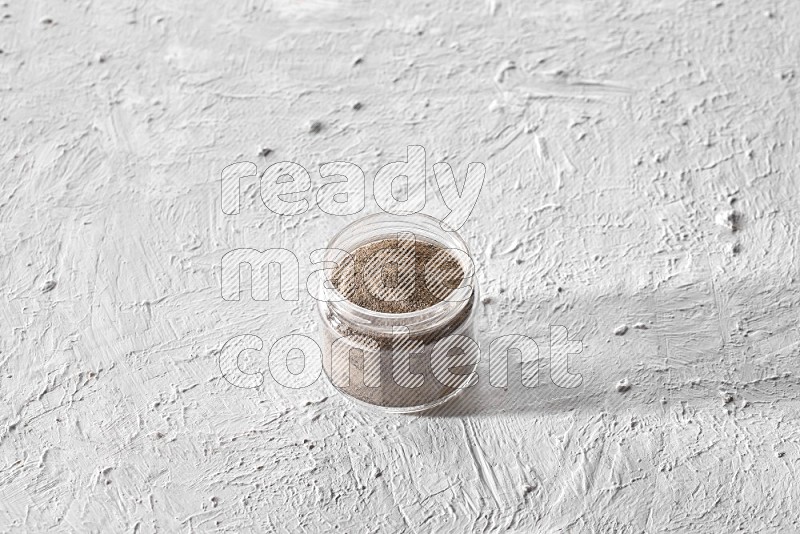 A glass jar full of black pepper powder on a textured white flooring