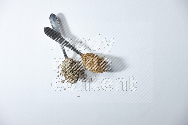 2 metal spoons full of cumin seeds and powder on white flooring