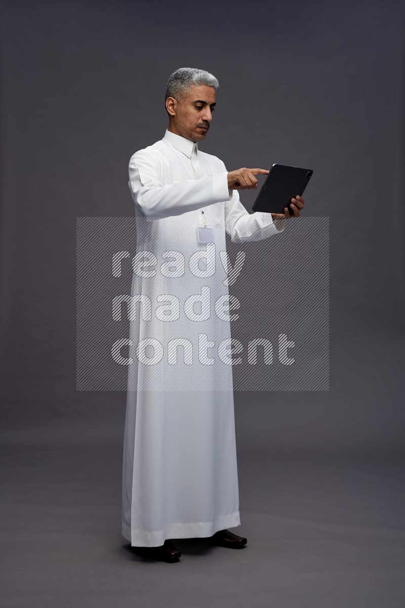 Saudi man wearing thob with neck strap employee badge standing working on tablet on gray background