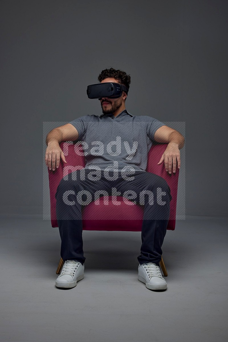 A man wearing casual and wearing VR while sitting on a burgundy chair eye level on a gray background