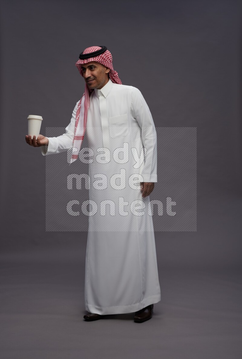 Saudi man wearing thob and shomag standing holding paper cup on gray background