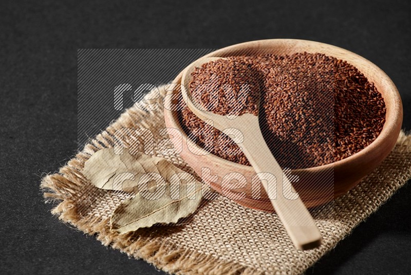 A wooden bowl full of garden cress seeds with wooden spoon full of the seeds on it on burlap fabric on a black flooring