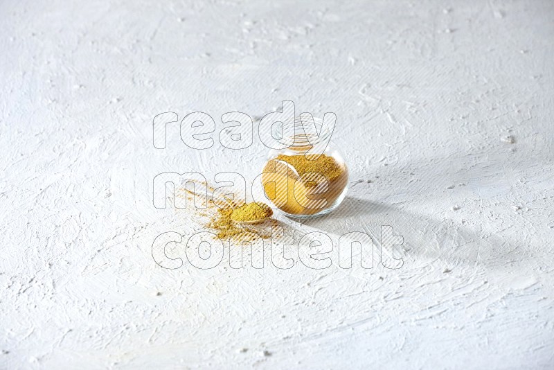 A glass spice jar and metal spoon full of turmeric powder on textured white flooring