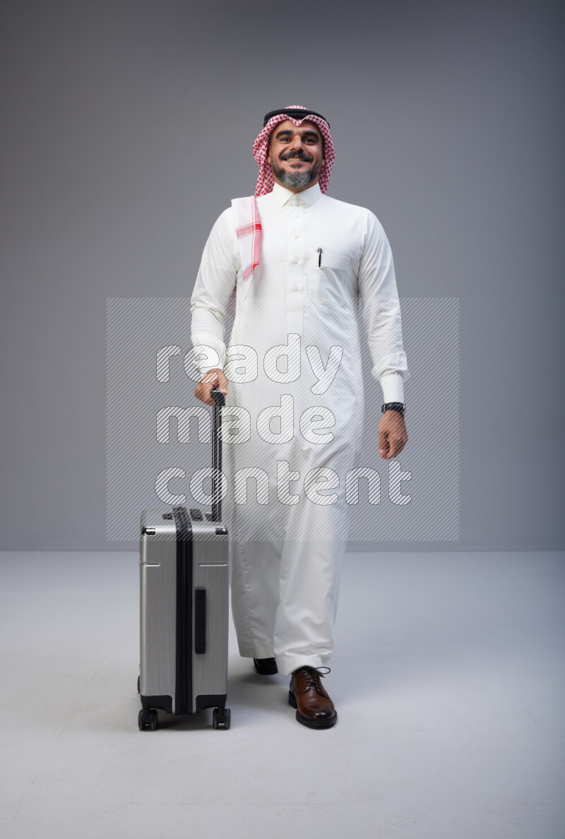 Saudi man wearing Thob and red Shomag standing holding Travel bag on Gray background