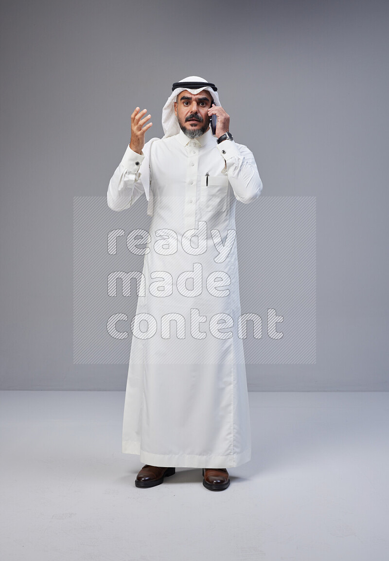 Saudi man Wearing Thob and white Shomag standing talking on phone on Gray background