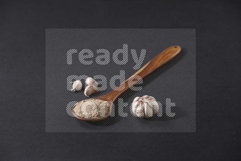 A wooden ladle full of garlic powder with a whole garlic bulb and some cloves beside it on a black flooring