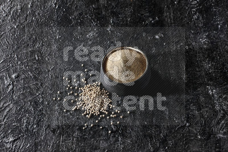 Black pottery bowl full of white pepper powder with white pepper beads on textured black flooring