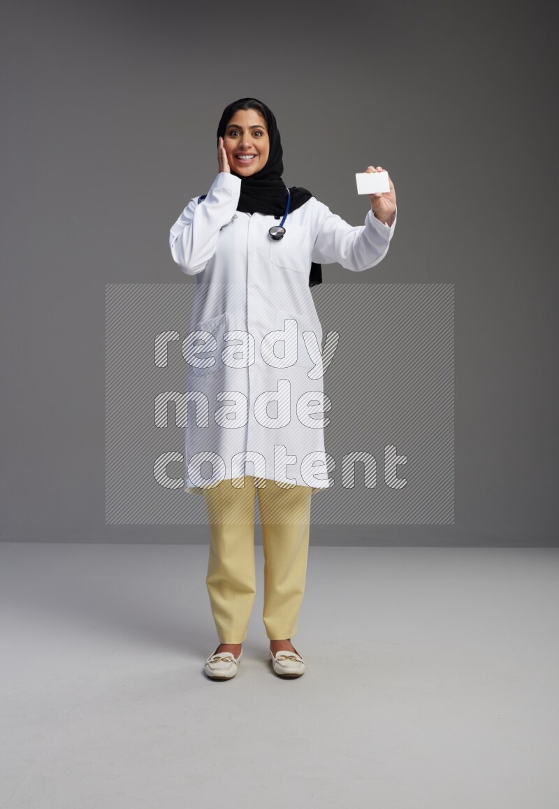 Saudi woman wearing lab coat with stethoscope standing holding ATM card on Gray background