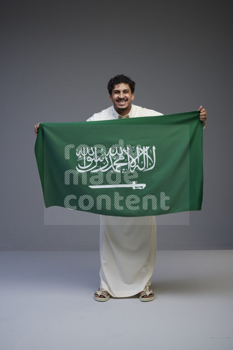 A Saudi man standing wearing thob holding big Saudi flag on gray background