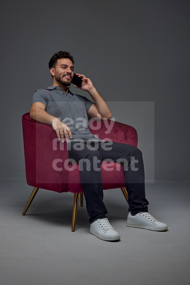 A man wearing casual and talking in his phone while setting on a burgundy chair eye level on a gray background