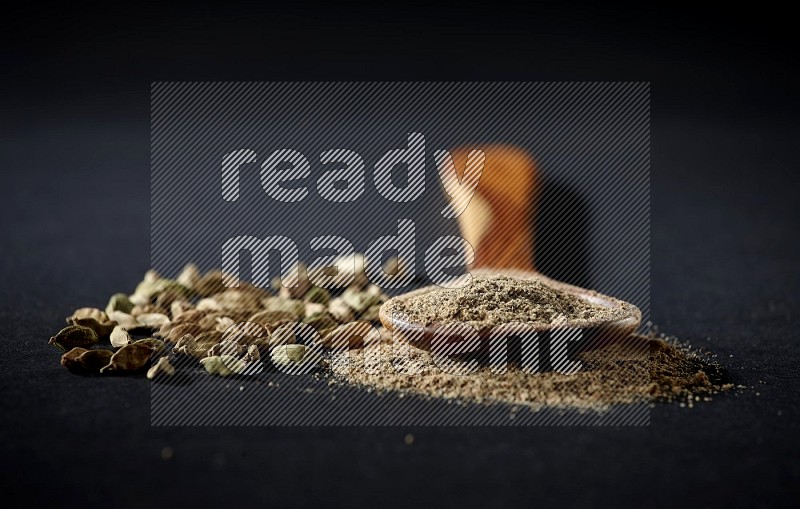 A wooden ladle full of cardamom powder and cardamom seeds beside it on black flooring